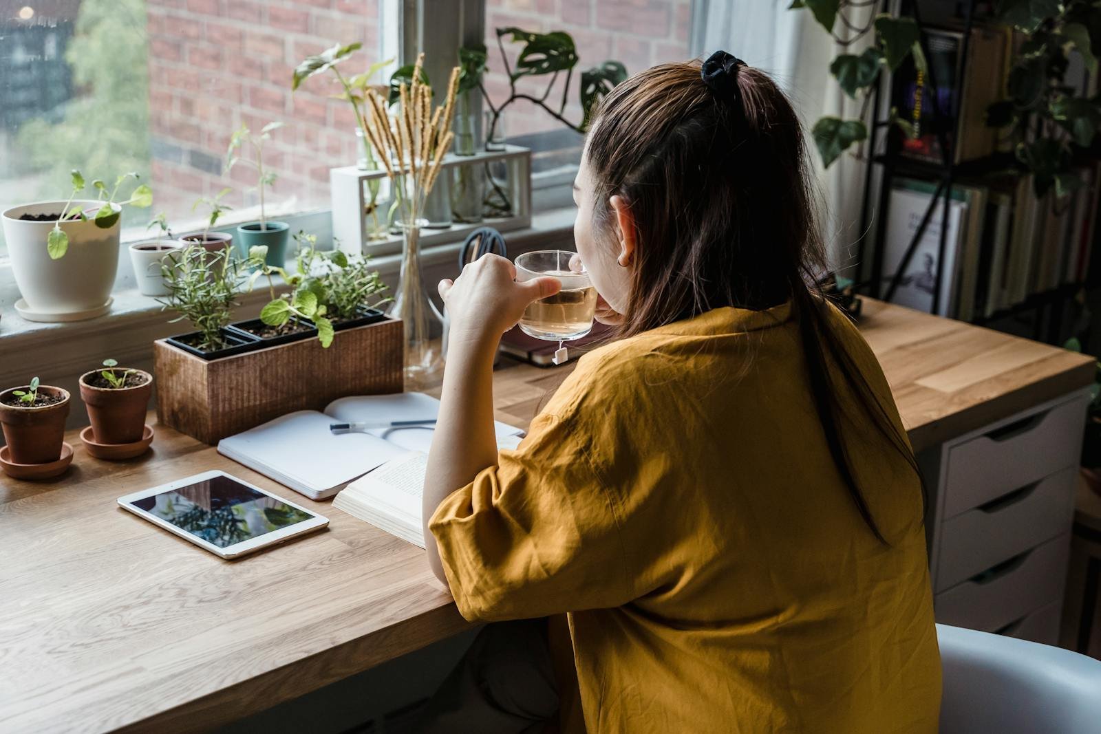 A woman sits at a desk drinking tea, surrounded by plants in a sunlit home office.